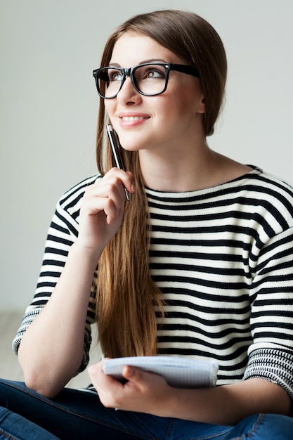 Solo ispirato. premurosa giovane donna in abiti a righe tenendo il blocco note e toccando il mento con la penna mentre è seduto sul pavimento di legno duro