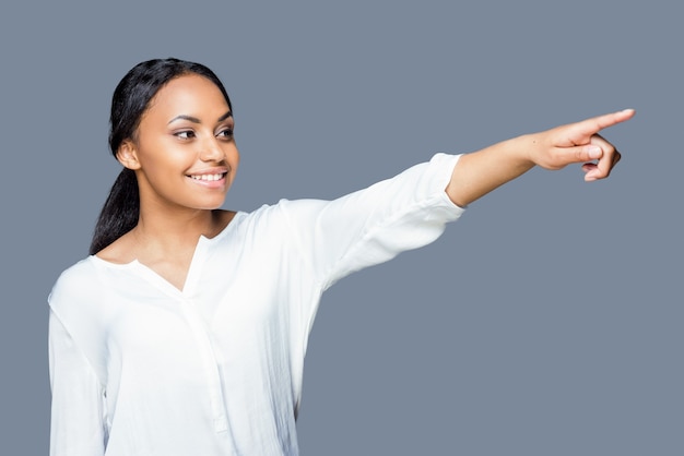 Just look at that! Attractive young African woman pointing away and smiling while standing against grey background