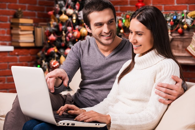 Just look at that! beautiful young couple using computer together while man pointing monitor and smiling with christmas decoration in the background