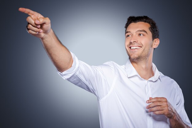 Just look at that! Happy young man in white shirt pointing away and smiling while standing against grey background