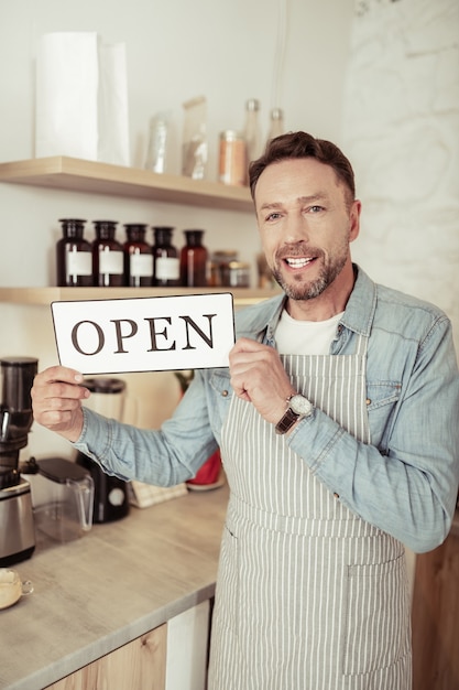 開店したばかりです。自分のコーヒーショップのキッチンに立っているハンサムな幸せな男は、その上に単語が開いている看板を持っています。