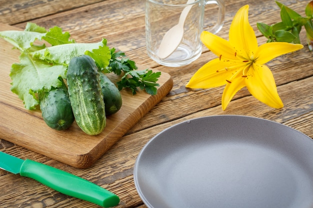 Just picked cucumbers and green salad on cutting board, knife, empty plate, glass cup and lily flower on old wooden boards. Just harvested vegetables. Top view.