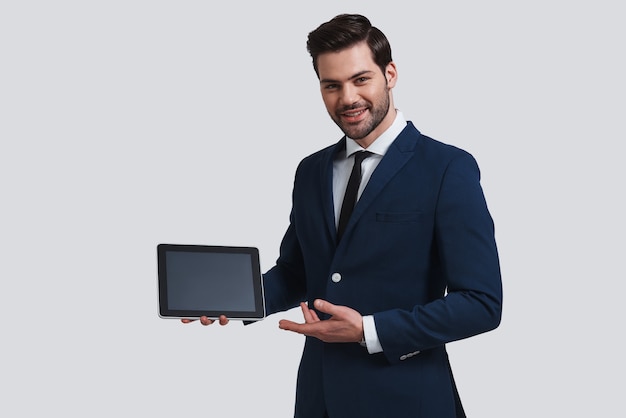 Just take a look. Good looking young man in full suit pointing at digital tablet and looking at camera with smile while standing against grey background