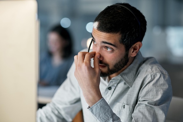 現代のオフィスで夜遅くにヘッドセットとコンピューターを使用しているときにストレスを感じている若い男のショットを時計で止められると思ったちょうどその時