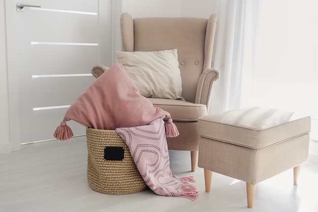 Un cesto di iuta fatto a mano con una piccola lavagna nera e cuscini e una coperta rosa sul pavimento accanto a una poltrona beige con un pouf su una superficie di carta da parati bianca. accogliente concetto di casa