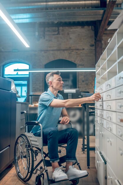 Kaart index. Geïnteresseerde serieuze man in blauwe t-shirt en jeans op rolstoel die archiefkastlade in kantoorruimte opent