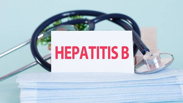 Kaart met woorden HEPATITIS B, stethoscoop, gezichtsmaskers en bloem op tafel op tafel. Persoonlijke beschermingsmiddelen op zachte blauwe achtergrond. Medisch en gezondheidszorgconcept.