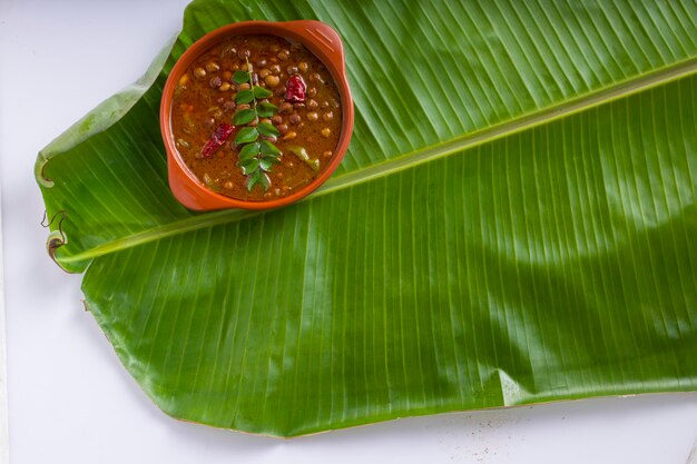 カダラカレーまたはチャナマサラカレーまたはひよこ豆カレーは、白い背景の新鮮な緑のバナナの葉の上にアテラコッタの陶器に配置されています。