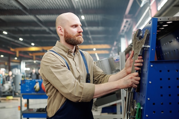 Kale bebaarde ingenieur in werkkleding enorme industriële moersleutel kiezen voor het uitvoeren van technisch werk in de fabriek