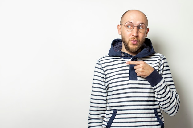 Kale jonge man met een baard in glazen van een jas met een capuchon wijst een vinger naar de zijkant op een afgelegen wit. gebaar van schok, verrassing