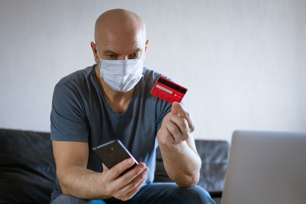 Kale man in een medisch masker met een creditcard zit op de bank op een laptop met een telefoon