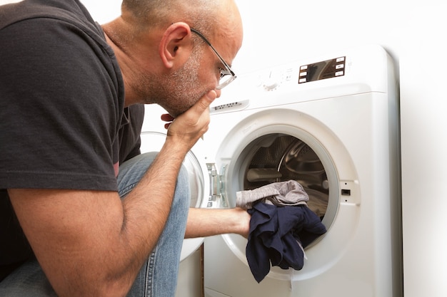 Kale man in grijze jeans en t-shirt zit thuis voor een wasmachine. laad de wasmachine met vuile kleren. lichte en ruime woonkamer met een modern interieur.