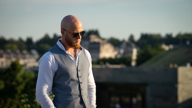 Kale man met een stijlvolle baard en zonnebril op een wazig stadsoppervlak tijdens zonsondergang