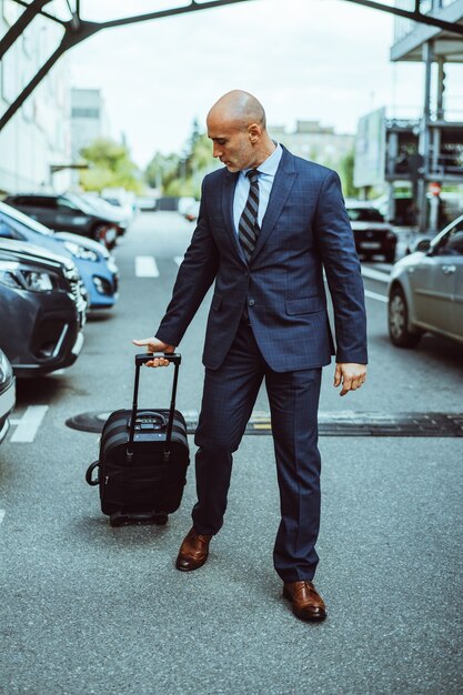 Kale zakenman lopen op de parkeerplaats van de luchthaven met koffer en geparkeerde auto 's