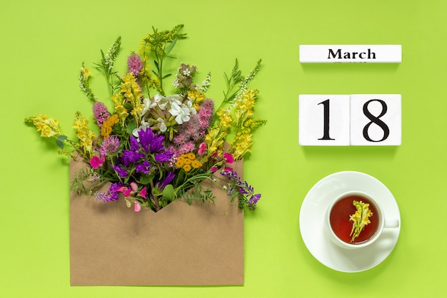 Kalender 18 maart. Kopje kruiden thee, kraft envelop met bloemen op groene achtergrond.