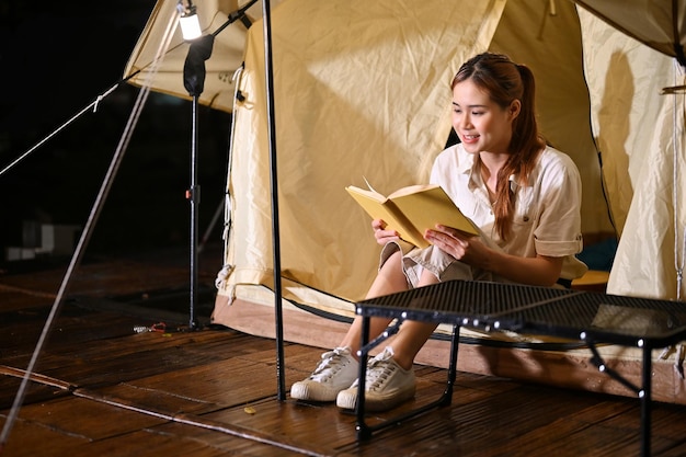 Kalme aziatische vrouw ontspant het lezen van een boek voor haar tent 's nachts kampeeractiviteit