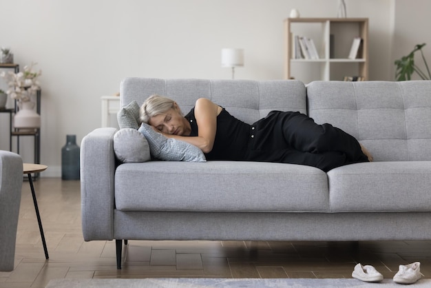 Kalme blonde vrouw van middelbare leeftijd, huiseigenaar, aan de zijkant liggende op een comfortabele bank, die overdag slaapt, zich moe voelt, uitgeput is, vermoeid is, ontspant, geniet van huishoudelijke vrije tijd.