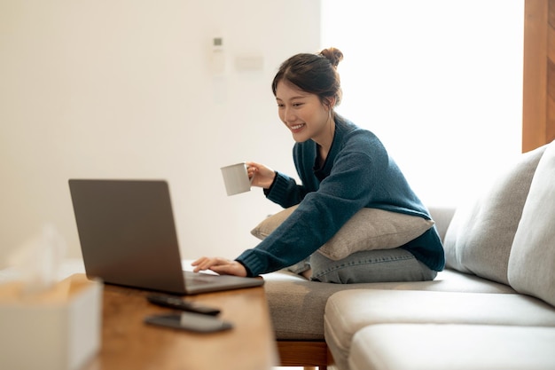 Kalme jonge Aziatische vrouw werkt bij laptop zittend op een gezellige bank gelukkig meisje surfen op internet of online winkelen tijdens zonnig weekend thuis ontspannen op de bank met computer