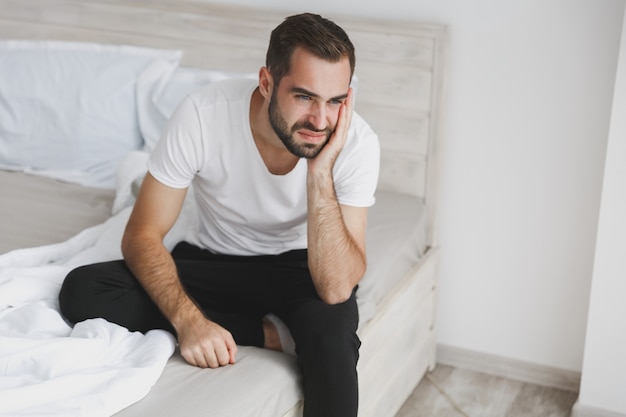 Kalme jonge knappe bebaarde man zittend in bed met witte laken kussen deken in slaapkamer thuis