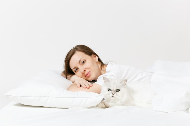 Kalme jonge vrouw liggend in bed met witte schattige Perzische zilveren chinchilla kat, laken, kussen, deken op witte muur