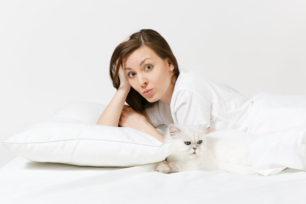 Kalme jonge vrouw liggend in bed met witte schattige Perzische zilveren chinchilla kat, laken, kussen, deken op witte muur