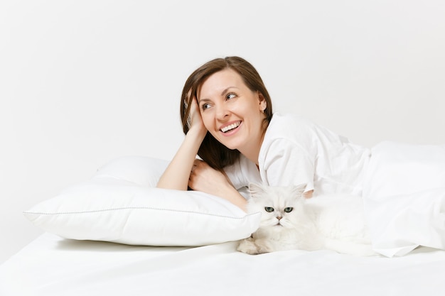 Kalme jonge vrouw liggend in bed met witte schattige Perzische zilveren chinchilla kat, laken, kussen, deken op witte muur