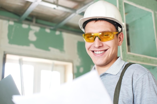 Kamer reparatie. Glimlachende jonge bouwer in helm en bril staat met een laptop en tekeningen in zijn handen tegen de achtergrond van een bouwplaats