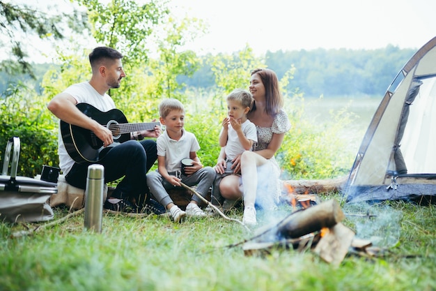 Kamperen aan een meer in het bos. gelukkige familie vader moeder en kleine kinderen zitten bij vuur en tent in de natuur. samen vrije tijd doorbrengen op vakantie. buitenshuis. Ouders met kinderen. vader gitaar spelen. Kamp