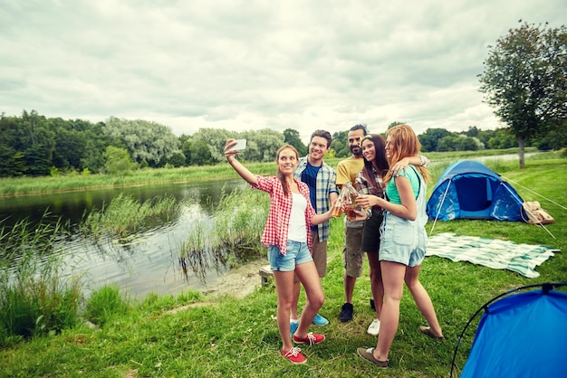 kamperen, reizen, toerisme, wandelen en mensenconcept - gelukkige vrienden met glazen flessen die cider of bier drinken en selfie nemen met een smartphone op de camping