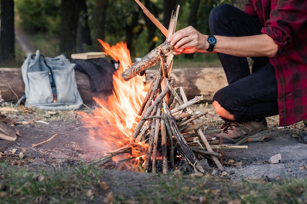 Kampvuur maken bij een bos. Going into the wild concept: kampeerplaats met vintage rugzak, thermoskan en man in vrijetijdskleding legt stukken hout in brand.