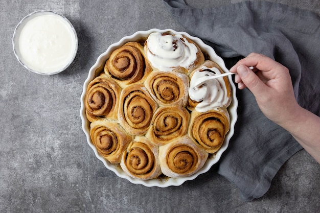 Kaneelbroodjes, broodjes in een ovenschaal met bruine suiker, gestremde roomsaus en kaneelstokjes met vrouwelijke hand decoreren broodjes met room.