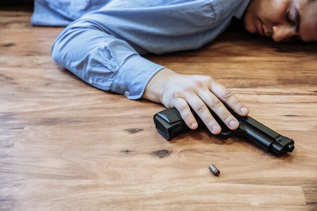 Kantoormedewerker ligt op de grond met een pistool in zijn handen. Zelfmoord door werkstress. Depressie of burn-out. Vreselijke levenssituatie. Man in de buurt van de laptop aan de balie.