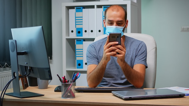 Kantoormedewerker met beschermend masker met behulp van telefoon typen zittend op modern kantoor tijdens coronavirus. Freelancer aan het werk op een nieuwe, normale werkplek, pratend pratend schrijvend met behulp van mobiele internettechnologie