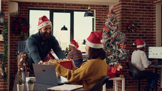 Kantoormedewerkers wisselen cadeautjes uit op het werk, voelen zich feestelijk met kerstmutsen op kantoor met kerstboom en versieringen. Vrouw ontvangt cadeau van collega tijdens seizoensvakantie.