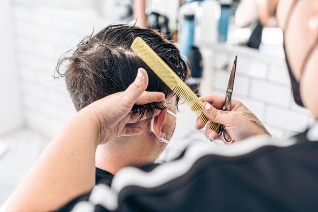 Kapper die een kam en schaar gebruikt om het haar van een jonge blanke man met masker in een salon te knippen