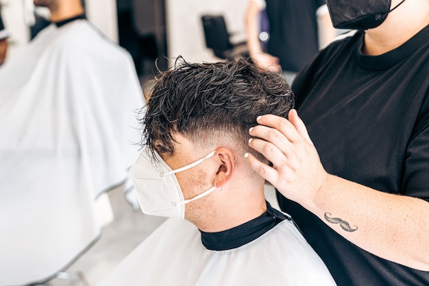 Kapper die het haar van een klant repareert met een masker zittend op een stoel in een kapperszaak