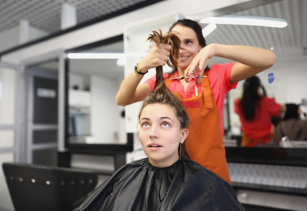 Kapper gaat een lok lang haar knippen met een schaar, vrouwelijke cliënt bang in de stoel van de meester