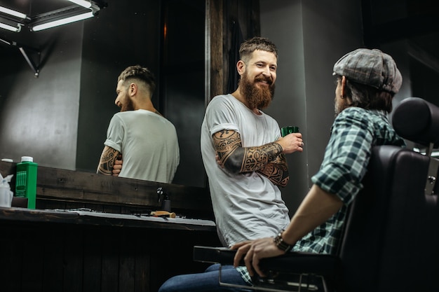 kapper met lange baard die glimlacht en een glas vasthoudt terwijl hij met zijn cliënt praat