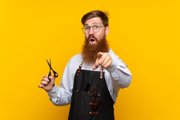 Kapper met lange baard in een schort over geïsoleerde gele muur verrast en wijzend voorzijde