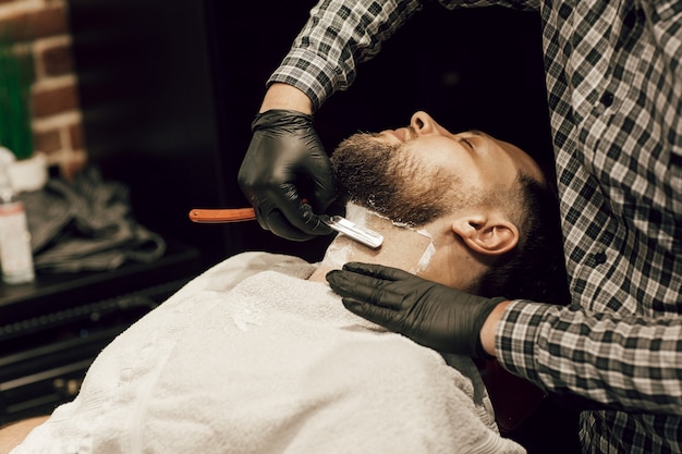 Foto kapper scheert een bebaarde man in een kapper. close-up van de handen van een kapper in zwarte handschoenen die de baard van een cliënt scheren.