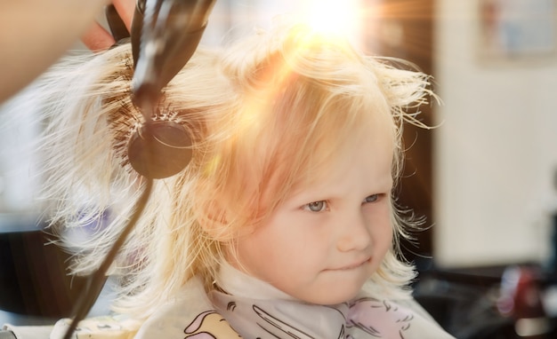 Kapper vrouw maakt modieus mooi kapsel voor schattig klein blond meisje kind in moderne kapperszaak, kapsalon. Kapper maakt kapsel voor jonge baby in kapperszaak. Concept kapsel en schoonheid