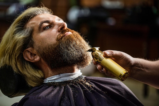 Kapper werkt met een baardtrimmer Hipster-cliënt krijgt kapsel Handen van een kapper met een baardtrimmer close-up Bebaarde man in de kapperszaak Man bezoekt haarstylist in de kapperszaak