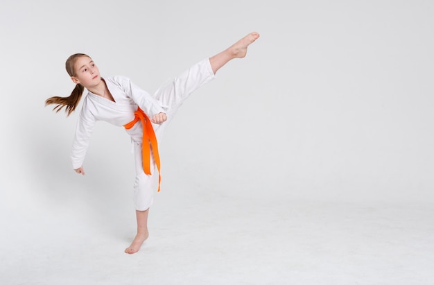 Karate girl at white studio background. sporty teenager in\
kimono doing kick. practicing martial arts. healthy lifestyle,\
active childhood and self defense concept, copy space