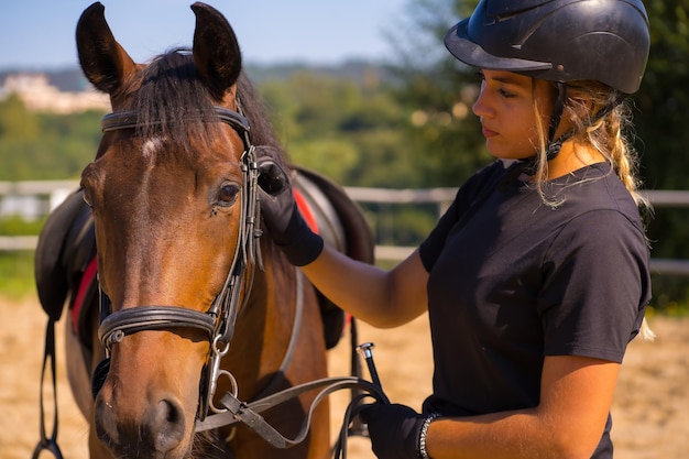 Kaukasisch blond meisje op een paard dat een bruin paard streelt en verwent, gekleed in een zwarte ruiter met veiligheidshoed