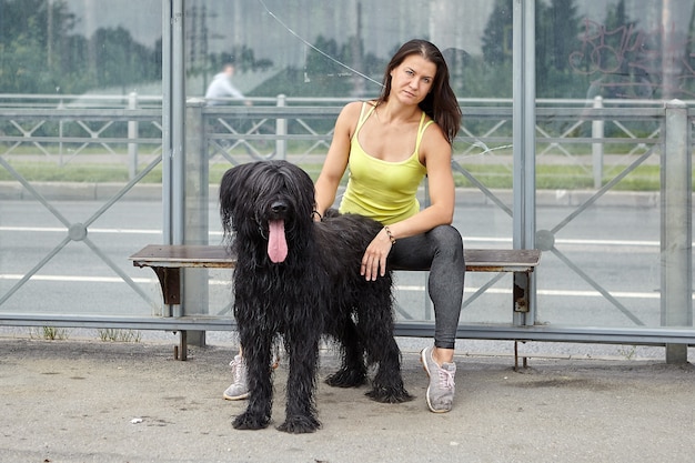 Kaukasisch ernstig meisje en haar zwarte ruige briard zitten op het station van het openbaar vervoer en wachten op de bus