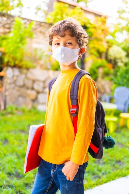 Kaukasisch kind met gezichtsmasker klaar voor terug naar school. Nieuwe normaliteit, sociale afstand, coronavirus-pandemie, covid-19. Oranje T-shirt, rugzak en een notitieblok in de hand