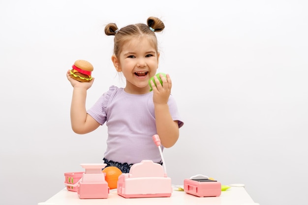 Kaukasisch lachend meisje met speelgoedhamburger en appel in de hand speelt in de winkel speelgoedkassa groenten mand schalen keuze tussen gezond en schadelijk voedselconcept