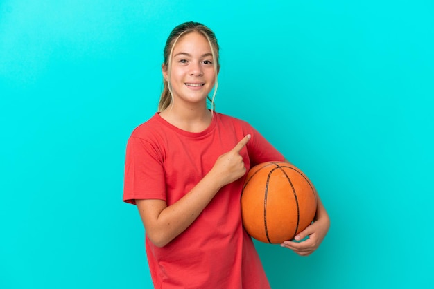 Kaukasisch meisje dat basketbal speelt geïsoleerd op een blauwe achtergrond en naar de zijkant wijst om een product te presenteren