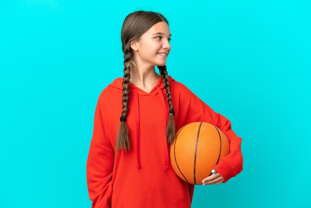 Kaukasisch meisje dat basketbal speelt geïsoleerd op een blauwe achtergrond op zoek naar de zijkant