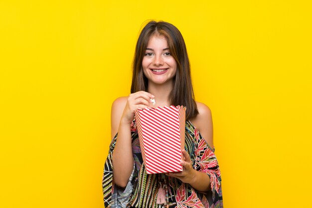 Kaukasisch meisje dat in kleurrijke kleding over geïsoleerde gele muur popcorns eet